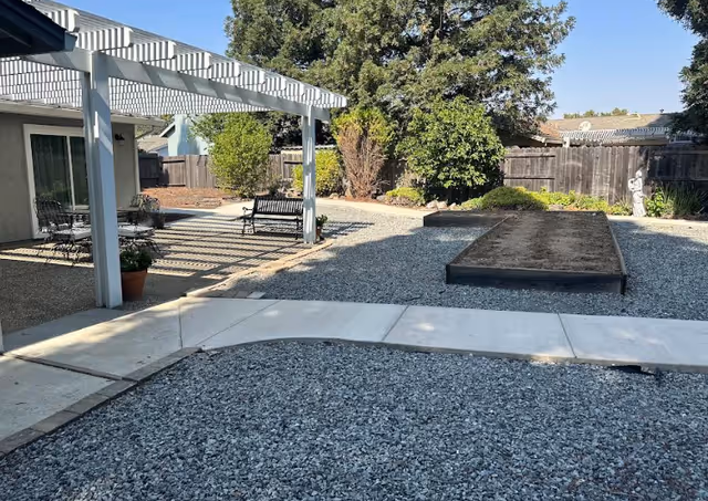 Outdoor garden area with gravel ground, a concrete pathway, raised garden beds, a wooden bench, patio chairs under a white pergola, and trees and shrubs along a wooden fence.