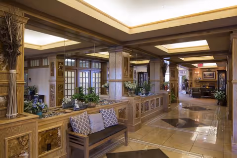Interior view of a senior living facility lobby or common area with polished tile floors, wooden decorative columns, a bench with cushions, plants, and a large mirror reflecting the space. The ceiling has recessed lighting and a coffered design.
