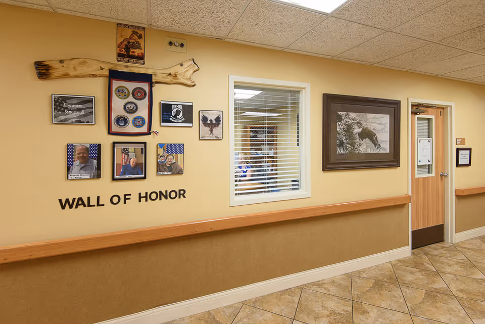 Interior hallway of a senior living facility with a 'Wall of Honor' display featuring photos and plaques honoring veterans. The wall is painted beige with a wooden handrail, and there is a window with blinds and a framed picture of an eagle on the adjacent wall. A wooden door with a small window and posted notices is visible at the end of the hallway.