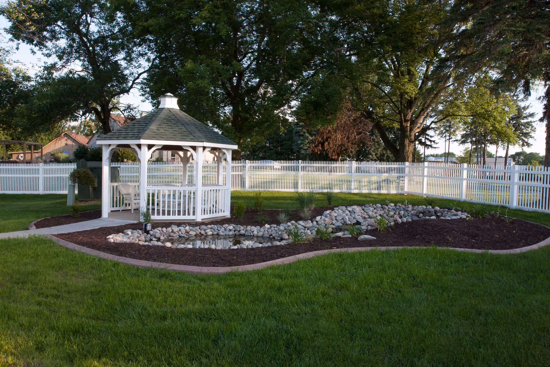 White wooden gazebo beside a small rock-lined pond in a grassy, white-fenced yard with large trees.