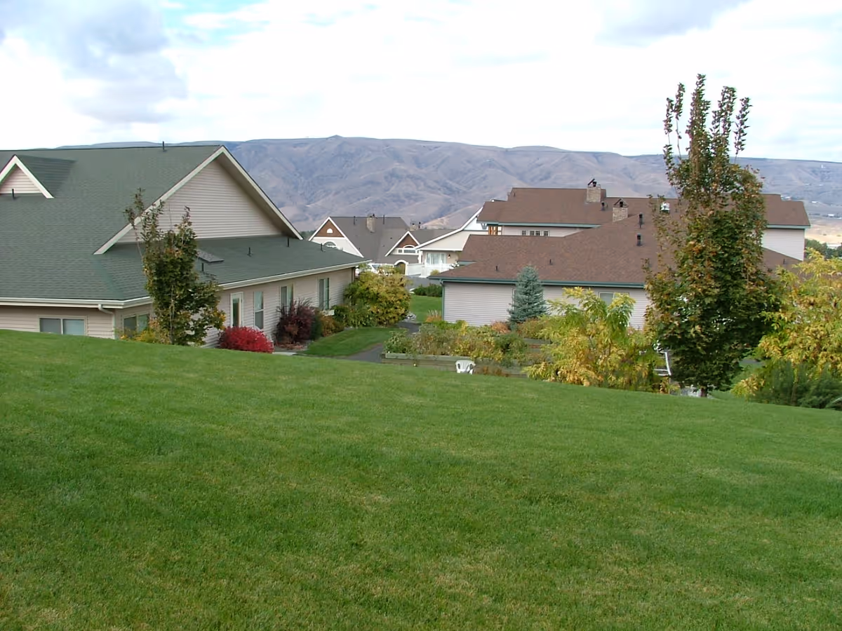 View of a senior living facility with multiple buildings featuring green and brown roofs, surrounded by well-maintained green lawns, trees, and shrubs, with hills visible in the background under a partly cloudy sky.
