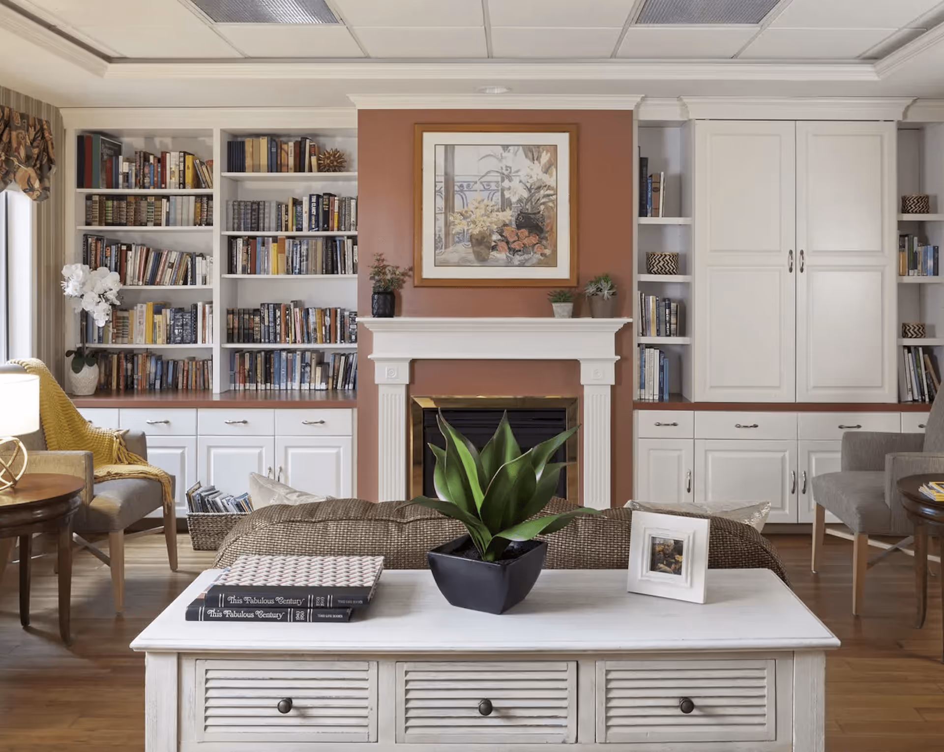 A cozy living room with built-in white bookshelves filled with books on either side of a white fireplace with a brown accent wall. A framed floral painting hangs above the fireplace. In front of the fireplace is a white coffee table with a green potted plant, two books, and a small framed photo. There are two armchairs with a yellow throw blanket on the left and a gray armchair on the right, with wooden side tables beside them. The floor is wooden, and the room has a warm, inviting atmosphere.