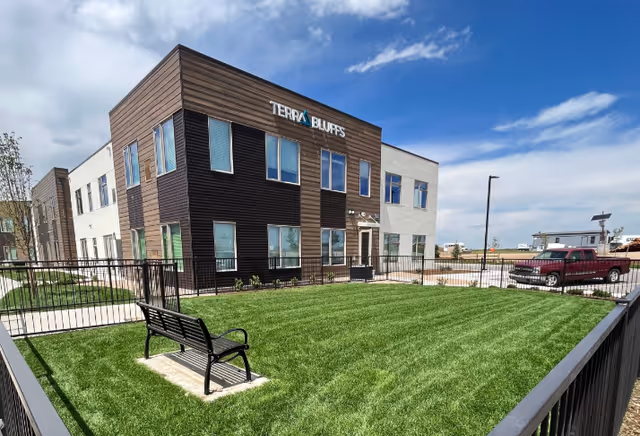 Exterior view of a two-story building named Terra Bluffs with a fenced grassy area and a black bench in the foreground. A red pickup truck is parked on the right side near the building under a partly cloudy sky.