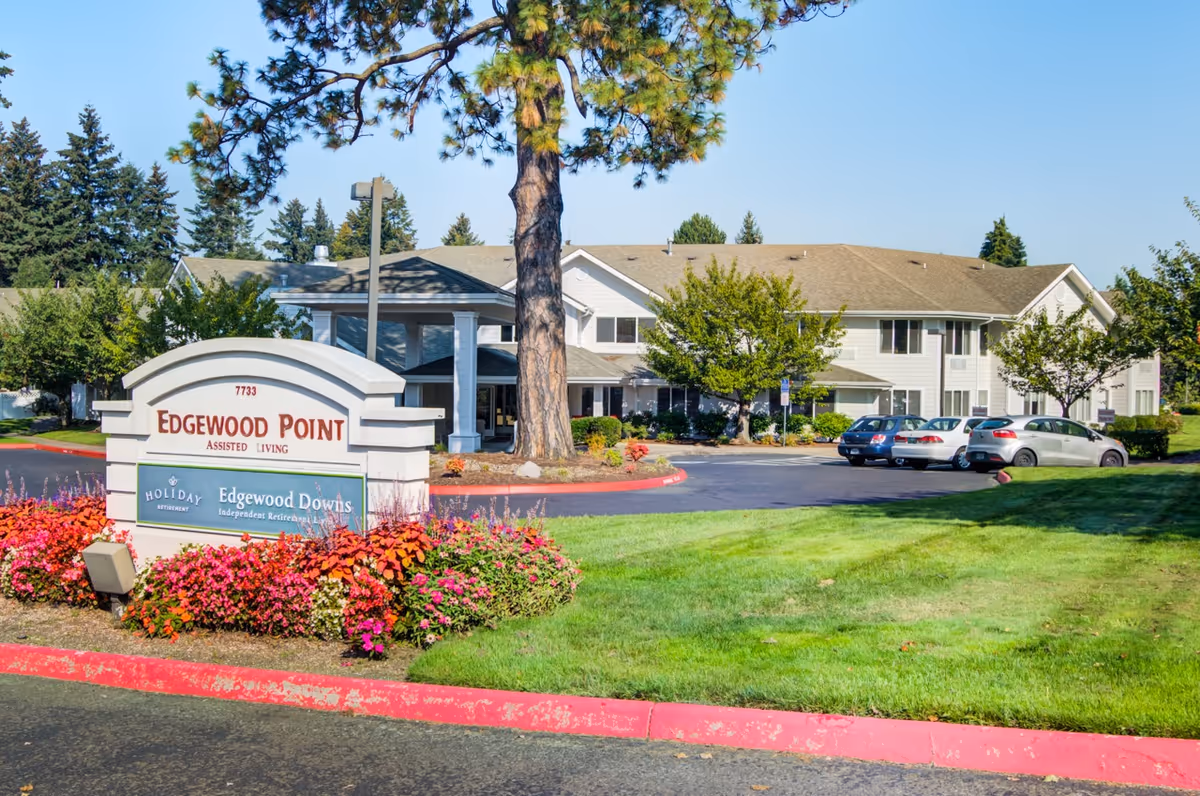 Exterior view of Edgewood Point Assisted Living facility with a large sign in front surrounded by colorful flowers and green grass. The building is two stories with white siding and a covered entrance. Several cars are parked in the parking lot, and trees are visible around the property under a clear blue sky.