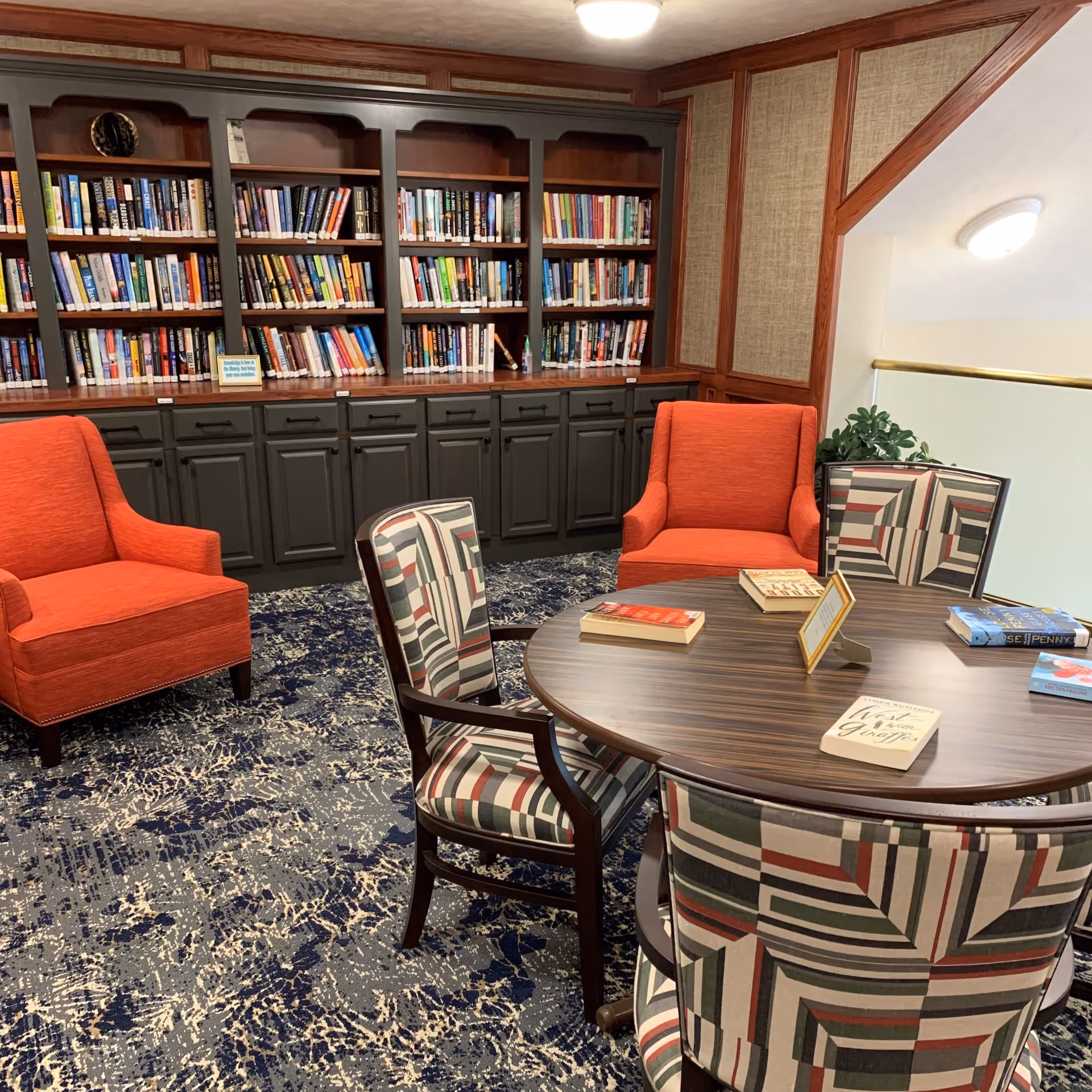 A cozy reading area in a retirement community featuring a round wooden table with several books on it, surrounded by four chairs with patterned upholstery and two orange armchairs. Behind the seating area is a large built-in bookshelf filled with books, set against a wall with wood paneling and textured wallpaper. The floor is covered with a blue and white patterned carpet.