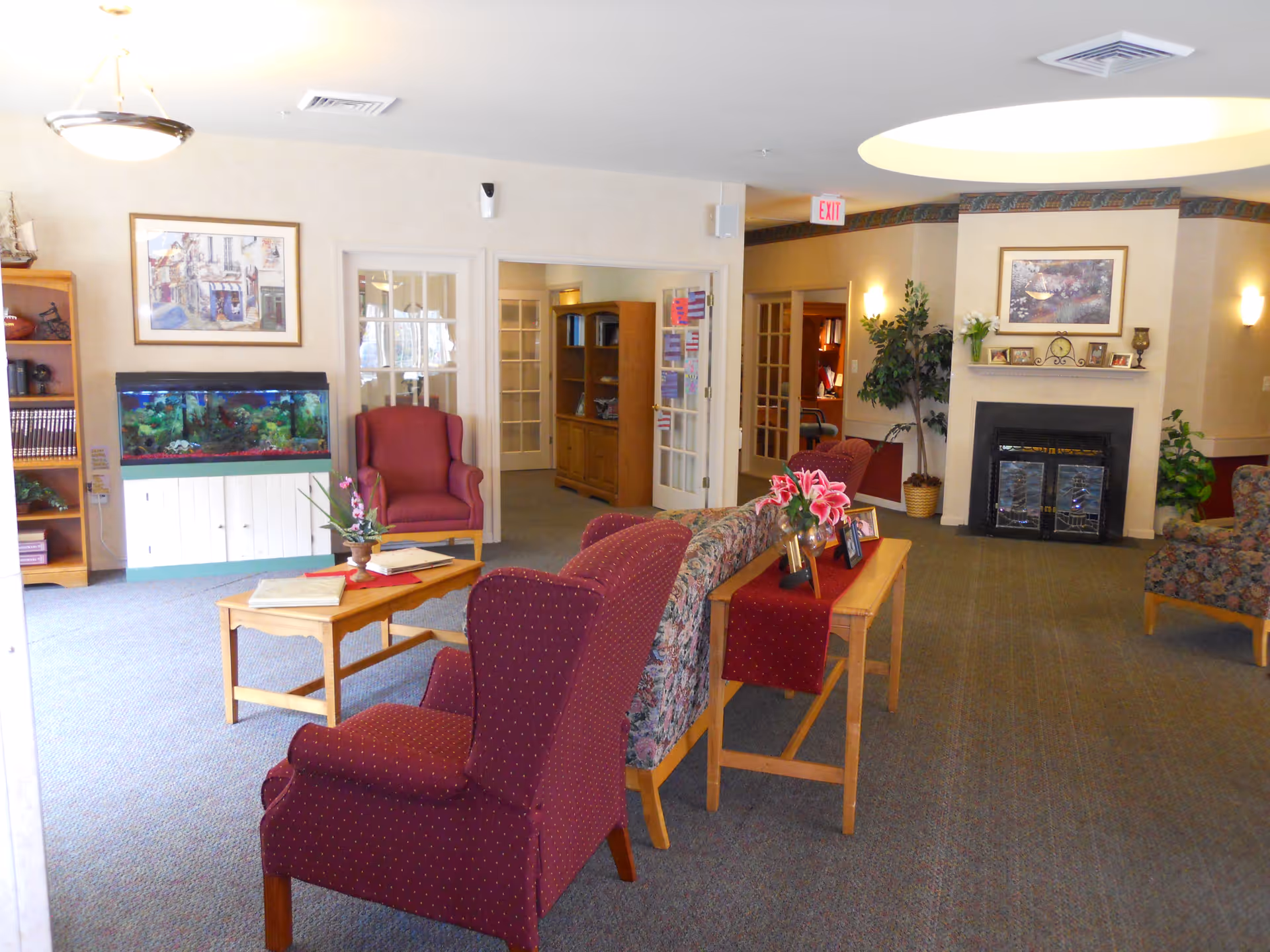 A cozy senior living community lounge area with upholstered armchairs and a floral-patterned sofa arranged around a wooden coffee table. There is a fish tank on a white cabinet against the wall, framed artwork, a fireplace with decorative items on the mantel, potted plants, and French doors leading to other rooms.