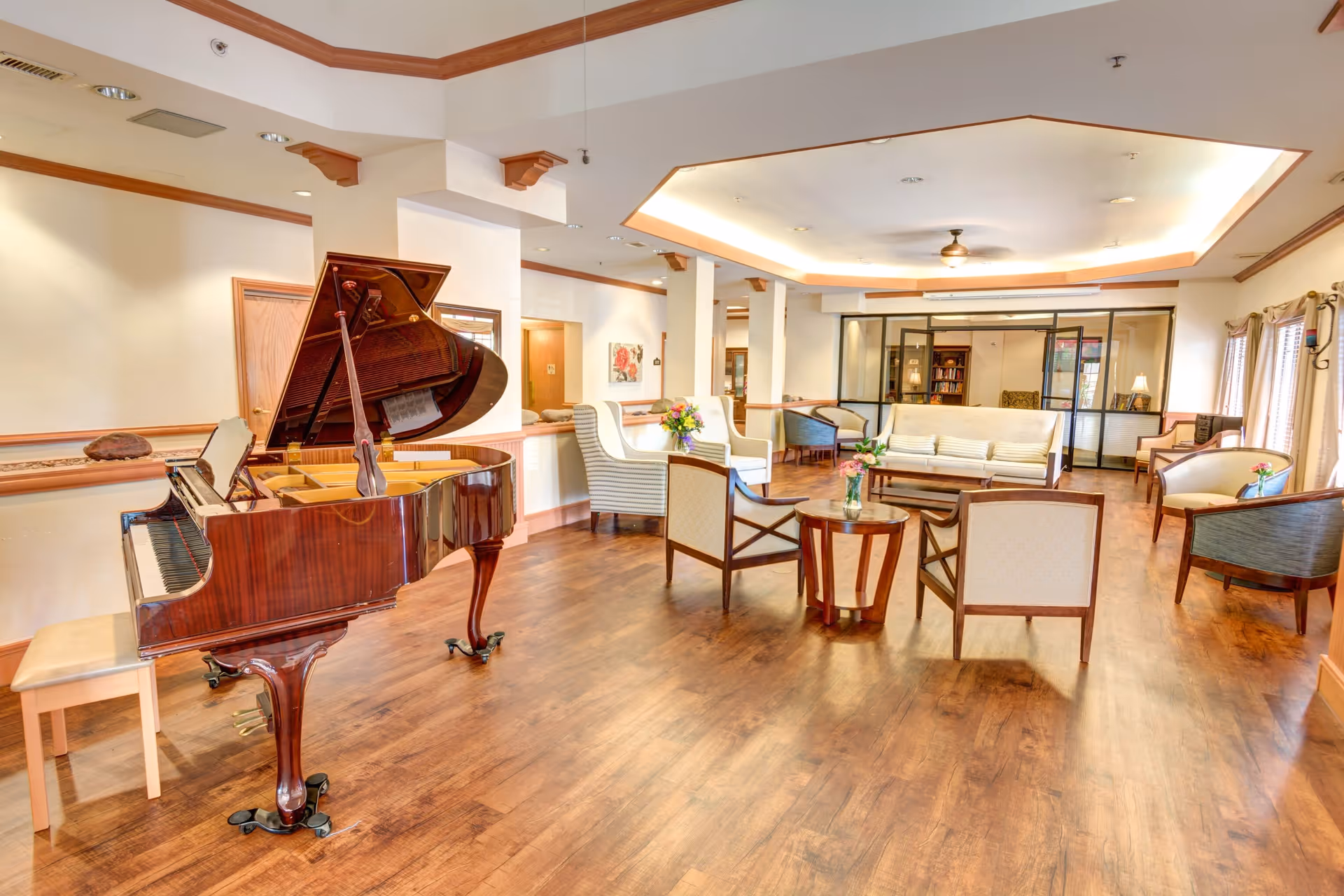 A spacious and well-lit common area with wooden flooring, featuring a grand piano on the left side and several seating arrangements including armchairs and sofas around small tables with flower vases. The ceiling has recessed lighting and a ceiling fan, and large windows with curtains allow natural light to enter the room.