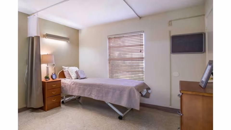 A simple nursing facility bedroom with a single bed, bedside table and lamp, window with blinds, and a dresser.
