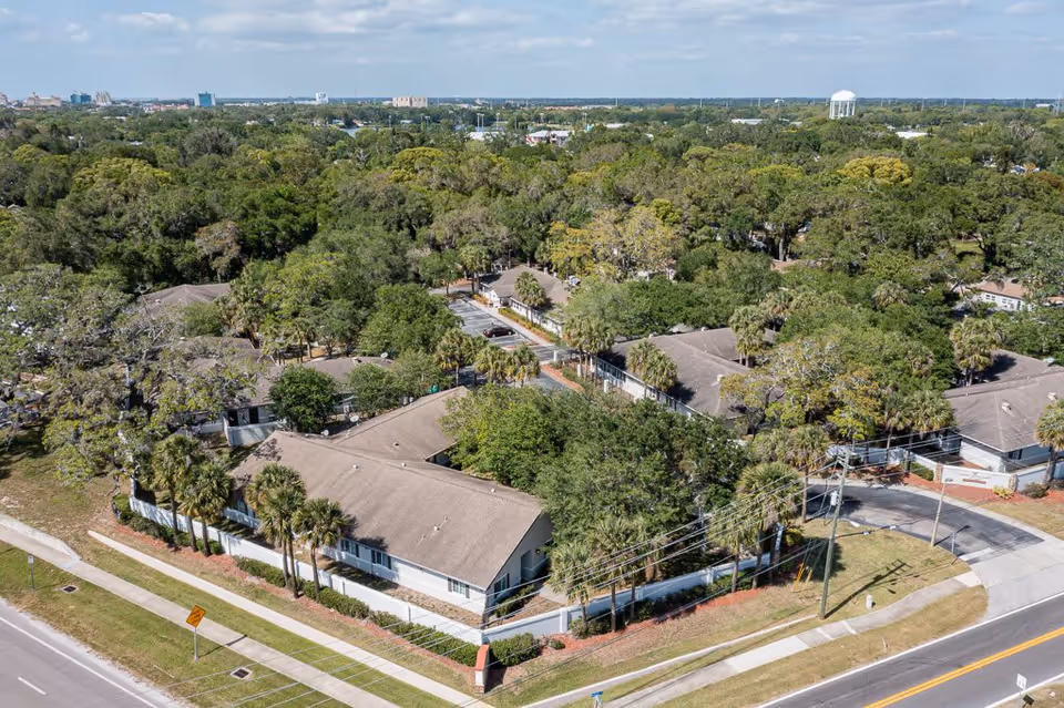 Aerial view of a senior living facility named Cottages at Belleair, showing multiple single-story buildings surrounded by trees and greenery, with roads and sidewalks bordering the property.