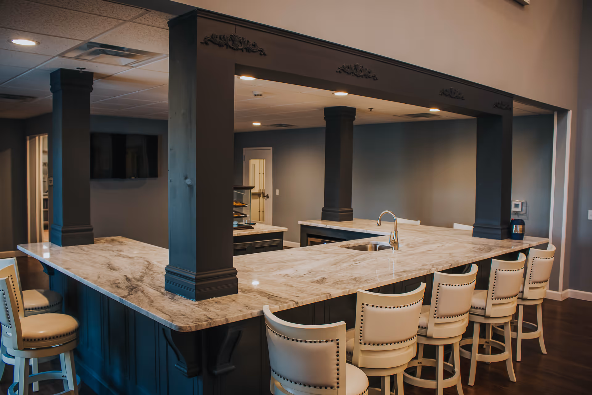 Interior view of a senior living facility's kitchen area featuring a large marble countertop island with a built-in sink and faucet. The island is surrounded by six cream-colored cushioned bar stools with nailhead trim. The space has dark pillars supporting a decorative overhead frame with recessed lighting. The walls are painted in a muted blue-gray color, and there is a door and a mounted TV in the background.