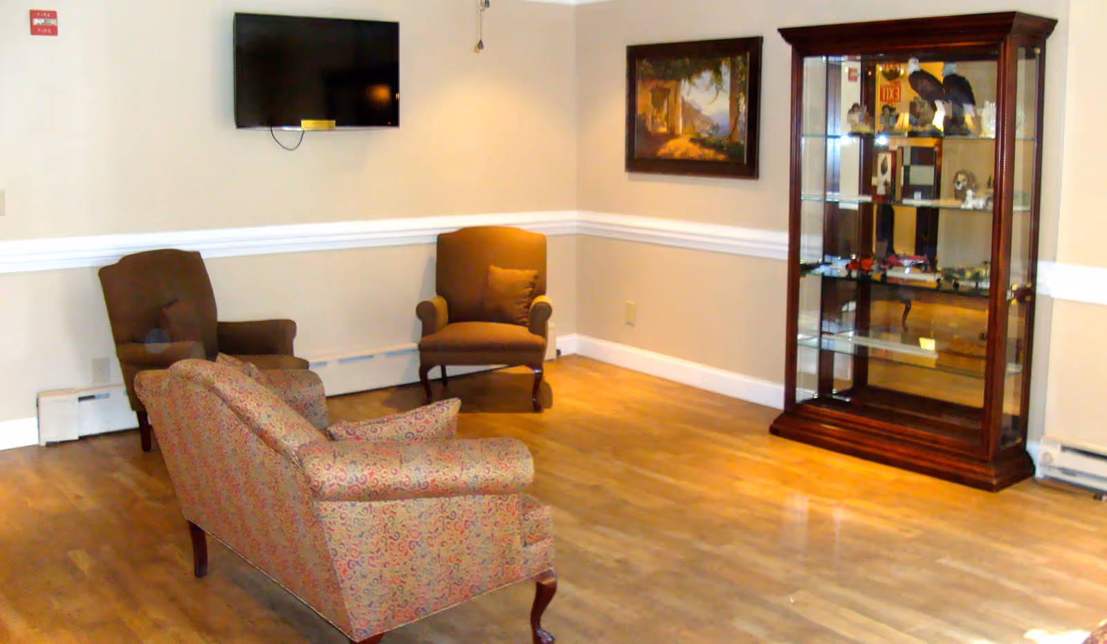 Small sitting room with upholstered chairs, a wall-mounted TV, and a glass display cabinet.