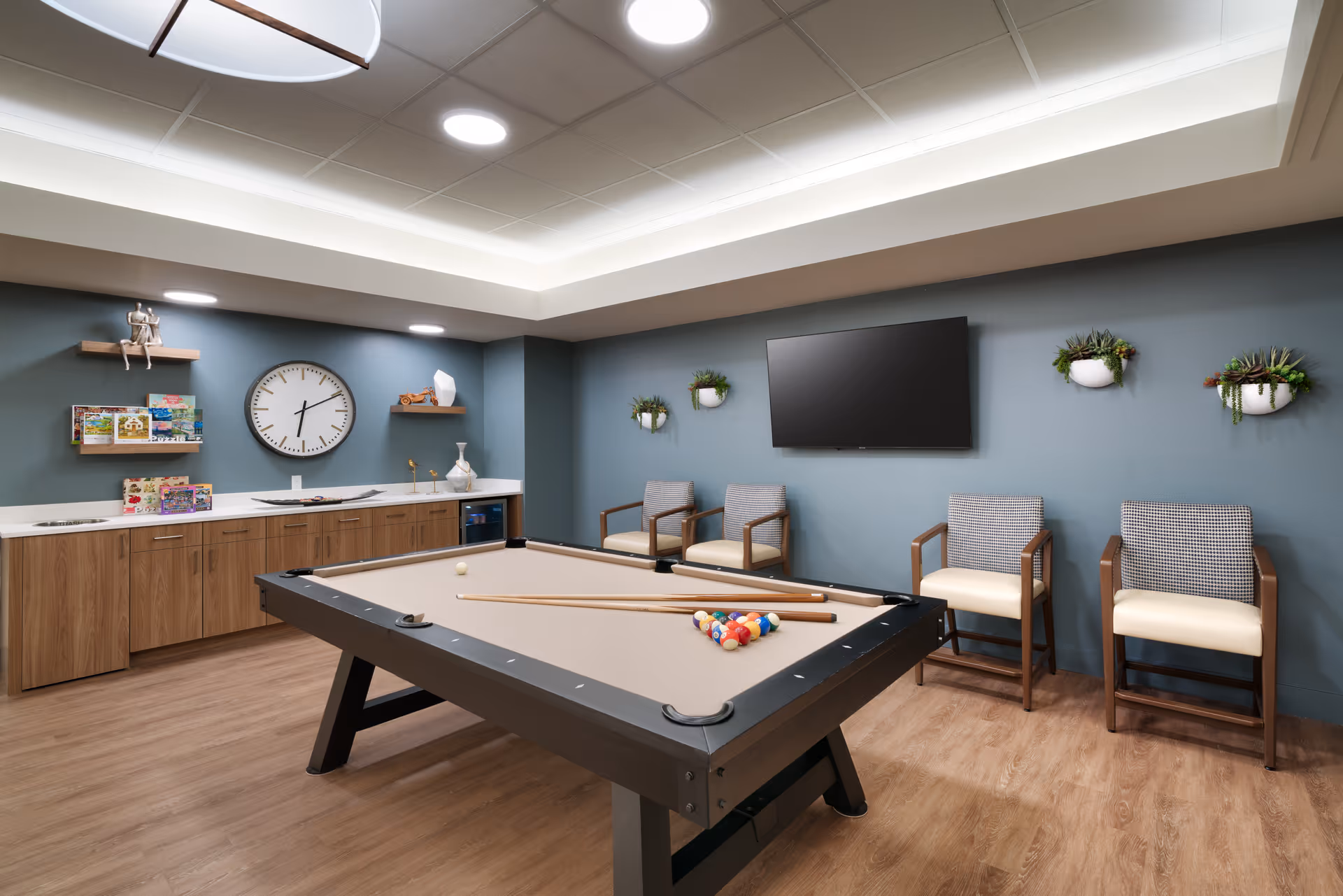 A recreational room with a pool table in the center, four wooden chairs with cushioned seats against a blue wall, three small wall-mounted planters with greenery, a large flat-screen TV mounted on the wall, and a wooden cabinet with shelves holding books and decorative items. The room has wood flooring and a ceiling with recessed lighting.
