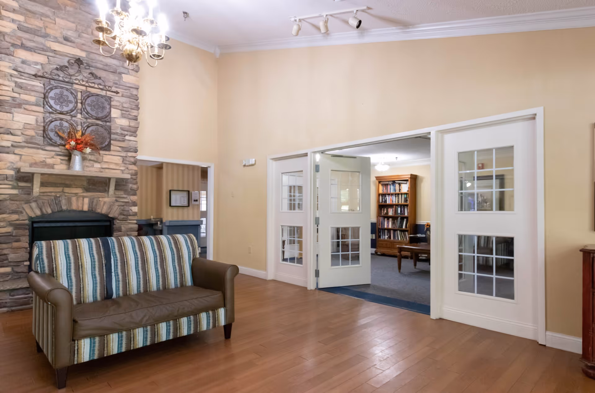 A cozy living room area with a striped and brown leather loveseat in front of a stone fireplace decorated with a metal wall art piece and a vase of flowers. The room has light yellow walls, wooden flooring, a chandelier, and white double doors with glass panes leading to a room with a wooden bookshelf and a coffee table.
