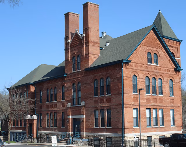 A large, historic red brick building with multiple tall chimneys and a steep green roof under a clear blue sky.