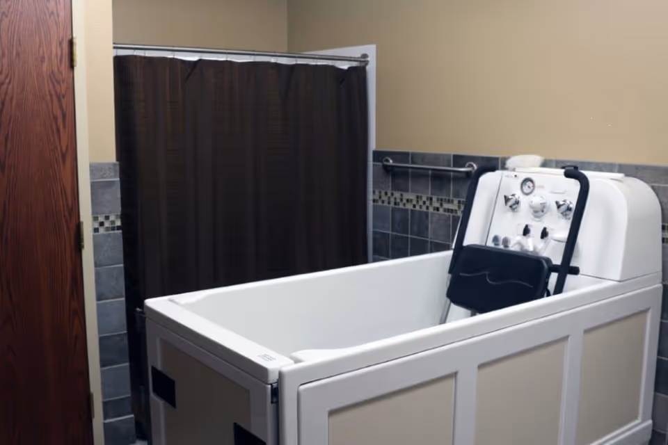 A bathroom with a specialized walk-in bathtub equipped with control knobs and a seat, a dark brown shower curtain, tiled walls with a decorative border, and a wooden door.