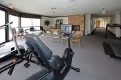 A spacious fitness room in an assisted living facility featuring exercise equipment including a recumbent bike and a treadmill. The room has large windows with blinds, a stone fireplace, chairs arranged in front of the fireplace, and a hallway leading to other areas.