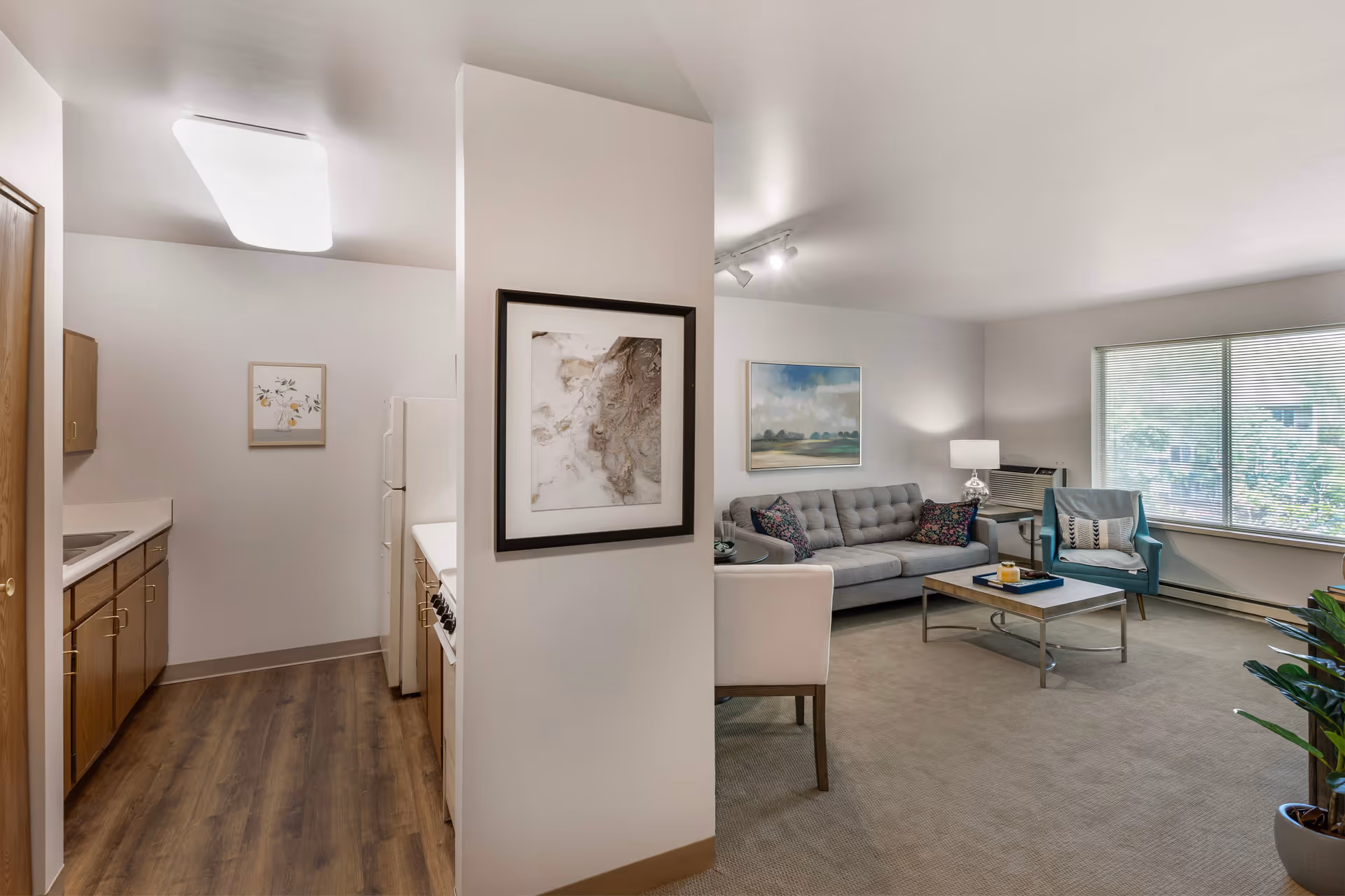 Interior view of a senior living facility showing a small kitchen area with wooden cabinets, a refrigerator, and a stove on the left, and a living room on the right with a gray sofa, a blue armchair, a coffee table, a side table with a lamp, a large window with blinds, and framed artwork on the walls.