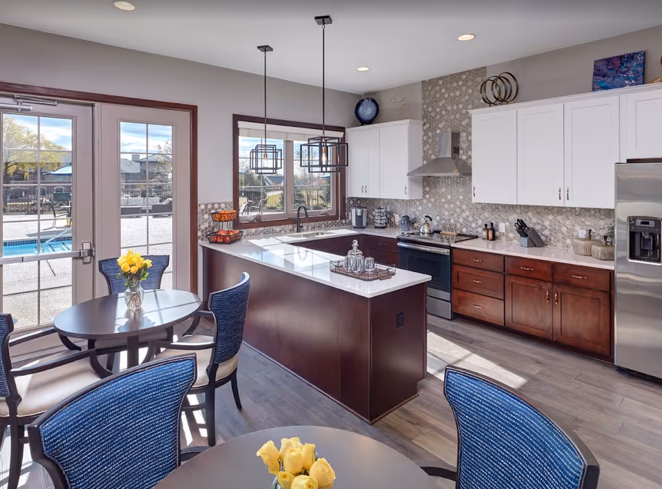 Modern kitchen with white upper cabinets and dark wood lower cabinets, a large island with a white countertop, stainless steel appliances including a refrigerator and stove, two pendant lights hanging above the island, and a dining area with round tables and blue cushioned chairs. Large windows and glass doors provide a view of an outdoor pool area.