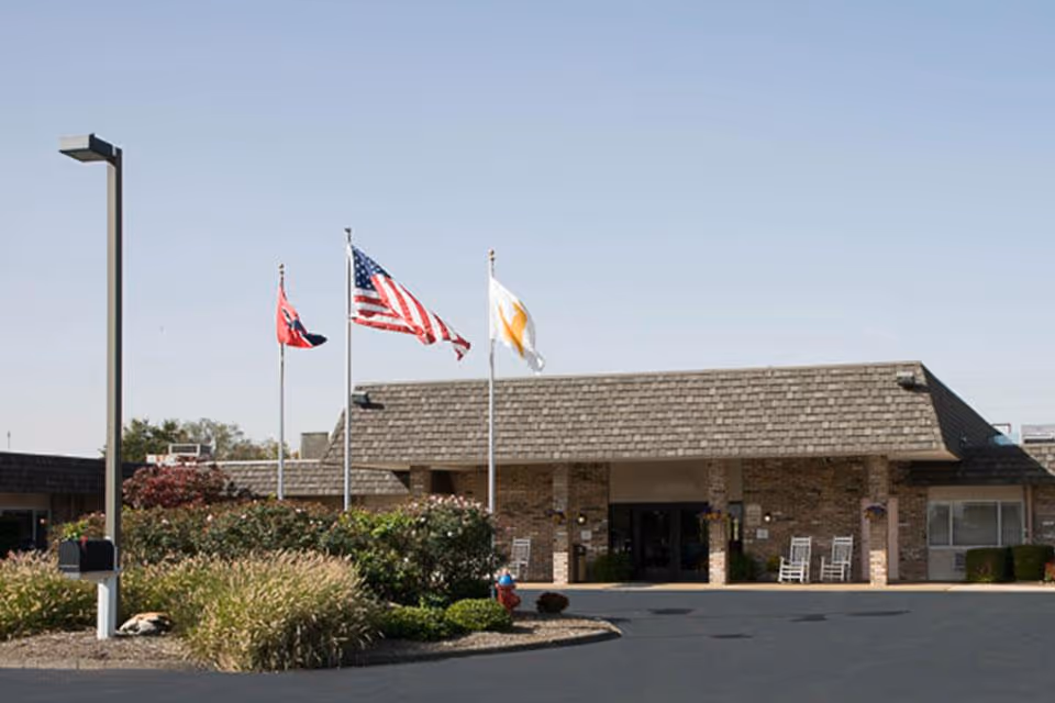 Front exterior view of a single-story brick building with a shingled roof, three flagpoles displaying the Tennessee state flag, the United States flag, and another flag, surrounded by landscaped bushes and plants under a clear sky.