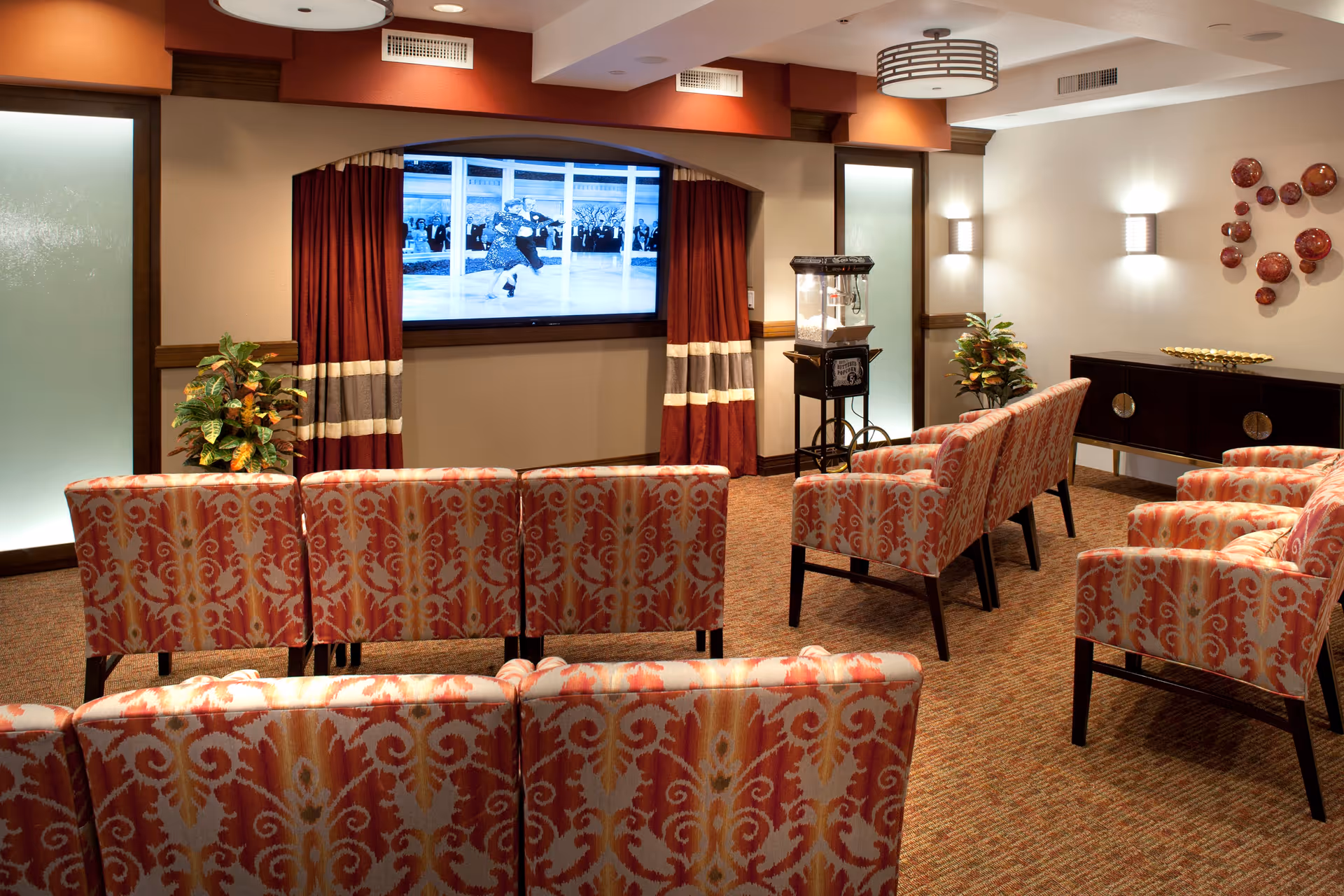 A cozy media room with patterned upholstered chairs arranged in rows facing a large flat-screen TV mounted on the wall. The TV screen shows a black and white image of a dancing couple. The room features warm lighting, red and beige curtains framing the TV, a popcorn machine on a stand, decorative plants, and a sideboard with decorative items on the right wall.