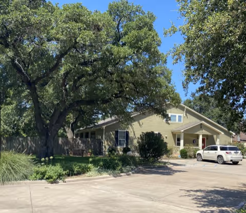Front exterior of Mercy House Waco, a single-story beige building with trees and a parked car.