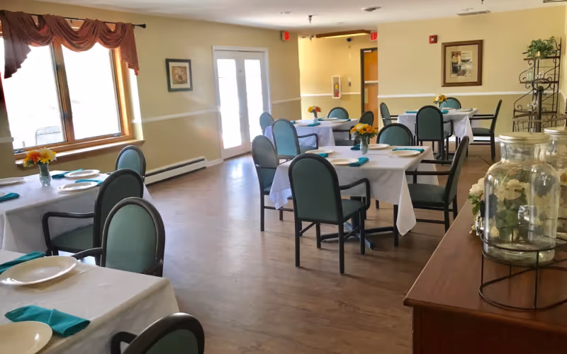 A bright dining room with several tables covered in white tablecloths, each set with plates and teal napkins. Green cushioned chairs surround the tables. Yellow flowers in small vases decorate each table. Large windows with red valances let in natural light, and a wooden sideboard with glass jars is visible on the right side of the room.