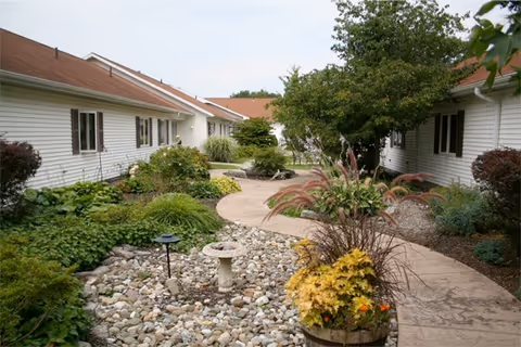 A landscaped outdoor courtyard area between single-story buildings with white siding and brown roofs. The courtyard features a curved paved walkway, decorative rocks, various green plants, shrubs, and a small birdbath or garden ornament. Trees provide some shade in the area.