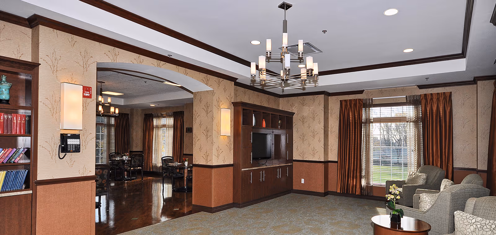 A cozy senior living facility common area with beige and brown wallpapered walls, a built-in wooden entertainment center with a TV, and a seating area with armchairs and a small round table with a flower vase. Large windows with brown curtains allow natural light to enter. In the background, there is a dining area with tables and chairs visible through an open archway.