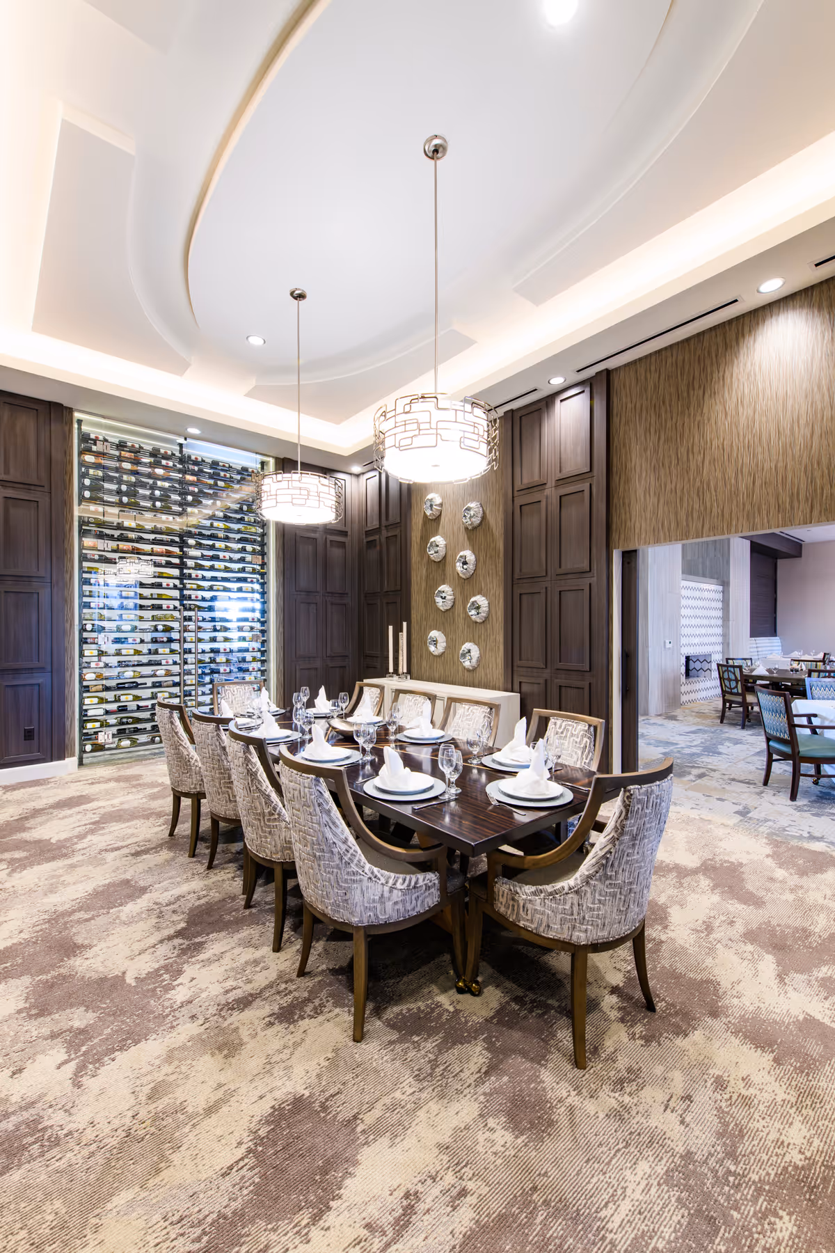 Elegant dining room with a long dark wooden table set for ten people with white napkins and glassware. The room features patterned upholstered chairs, a textured carpet, dark wood paneling, decorative wall plates, and two modern pendant lights hanging from a recessed ceiling. A glass-enclosed wine rack is visible in the background.