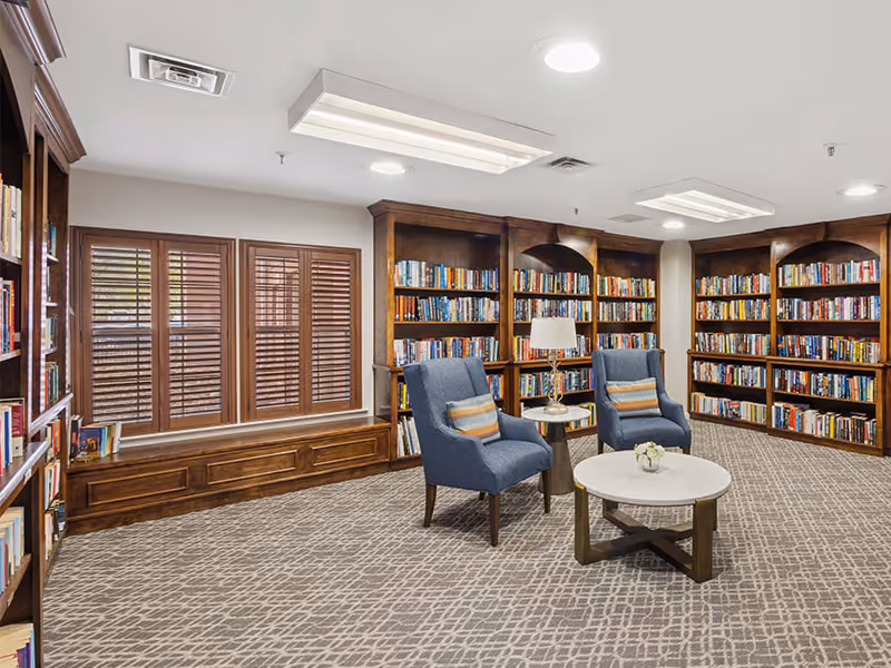 A cozy library room with wooden bookshelves filled with books lining the walls. Two blue upholstered armchairs with striped cushions are positioned around a small round table with a lamp and a flower vase. The room has a patterned carpet and windows with wooden shutters.