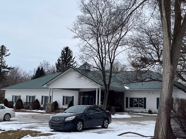 One-story white assisted living building with a green metal roof, bare trees, and cars parked on a snowy driveway.