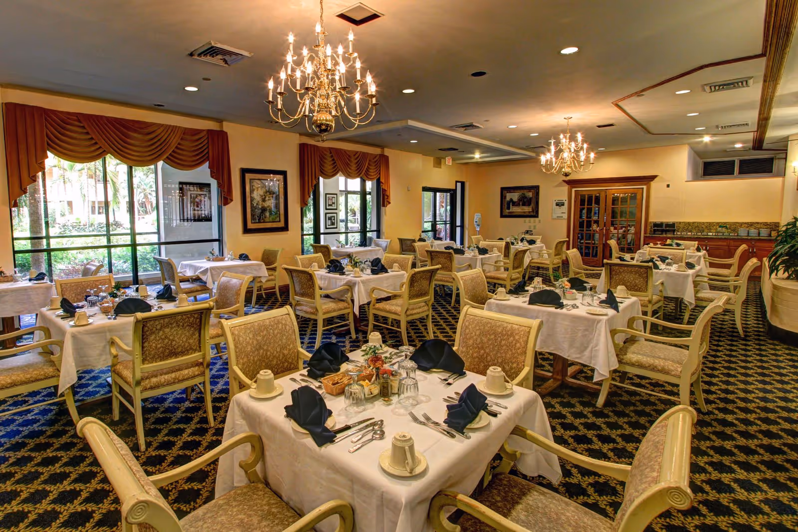 Formal dining room with multiple tables set with white tablecloths, upholstered chairs, and chandeliers.