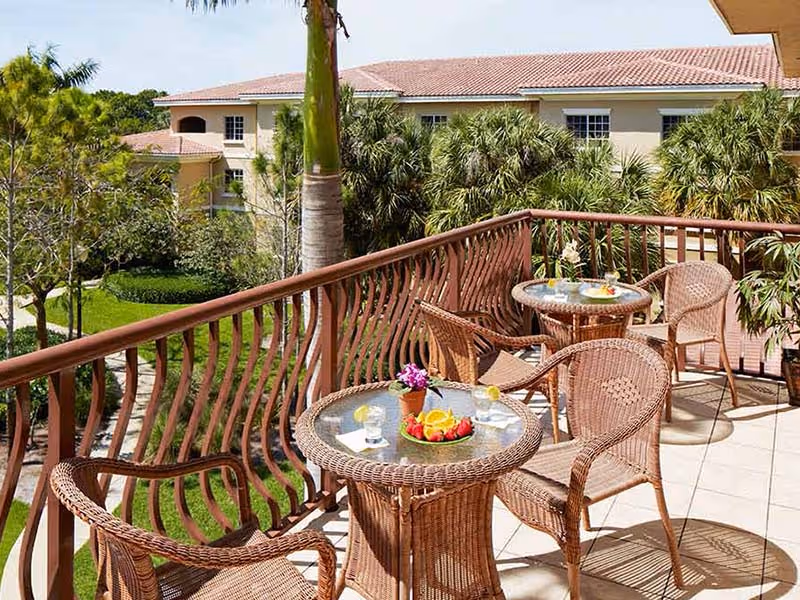 Outdoor balcony area with wicker tables and chairs. Each table has glasses of water with lemon slices and a plate of fruit. The balcony overlooks lush greenery and palm trees with a building in the background under a clear sky.
