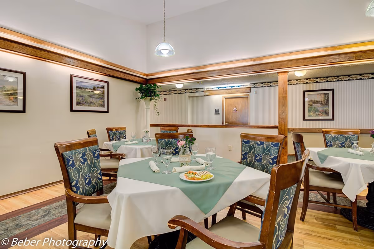 A dining room in a senior living facility with tables covered in white and green tablecloths, set with glassware, napkins, and a plate of food. The room features wooden chairs with patterned cushions, framed landscape paintings on the walls, and a hanging light fixture.