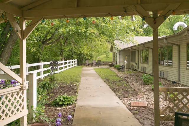 View of a covered patio area leading to a concrete pathway surrounded by garden beds with plants and flowers, a white fence on the left, and a single-story building on the right. At the end of the pathway, there is outdoor seating with a table and chairs under trees.