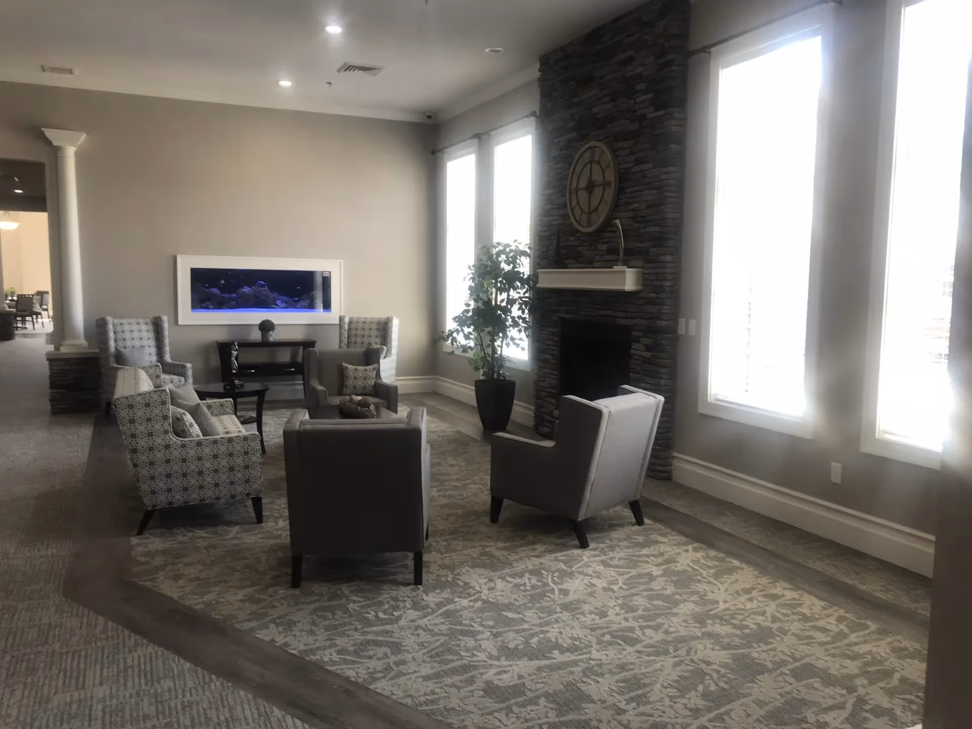 A senior living facility common area with several upholstered armchairs arranged around a small table on a patterned rug. The room features large windows letting in natural light, a stone fireplace with a large clock above it, and a built-in aquarium on the far wall. There is a potted plant near the windows and a hallway leading to another room in the background.