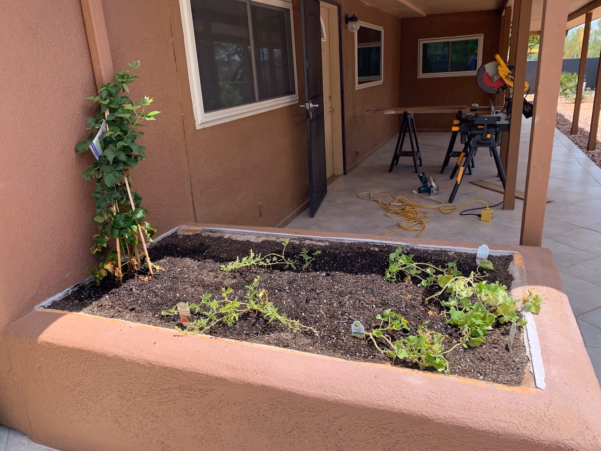 A raised garden bed with soil and small plants growing, located on a covered patio area with brown stucco walls. In the background, there is a door, windows, and some construction tools including a saw and extension cords on the tiled floor.