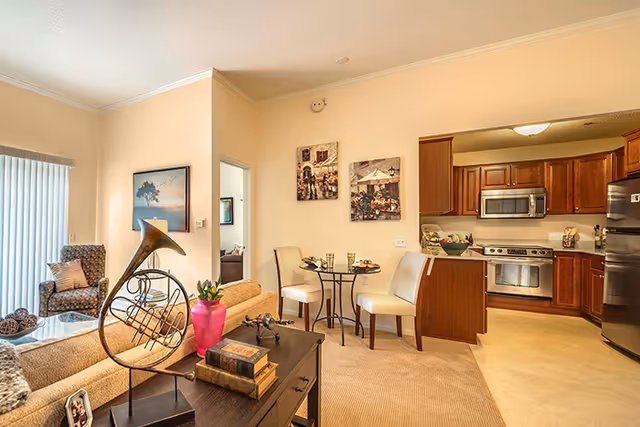 A cozy living area in a senior living facility featuring a beige sofa with a decorative French horn sculpture on a side table, a small round dining table with two chairs set for a meal, and a kitchen with wooden cabinets, stainless steel appliances including a microwave, stove, and refrigerator. The room has light-colored walls, carpeted flooring in the living area, and tile flooring in the kitchen. Artwork hangs on the walls and a sliding glass door with vertical blinds is visible.