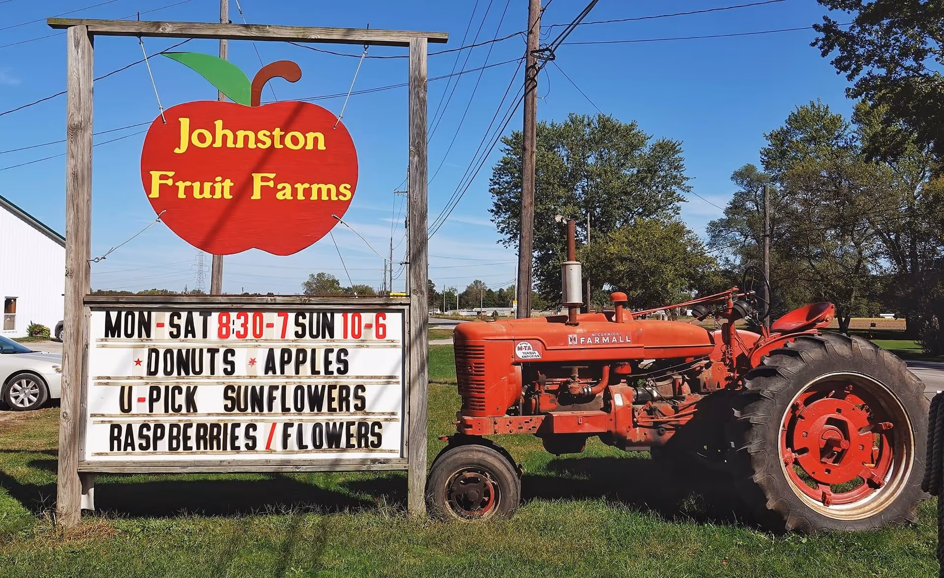 A wooden sign for Johnston Fruit Farms with a large red apple graphic at the top, displaying hours and offerings such as donuts, apples, u-pick sunflowers, raspberries, and flowers. Next to the sign is a vintage red Farmall tractor on grass with trees and a clear blue sky in the background.