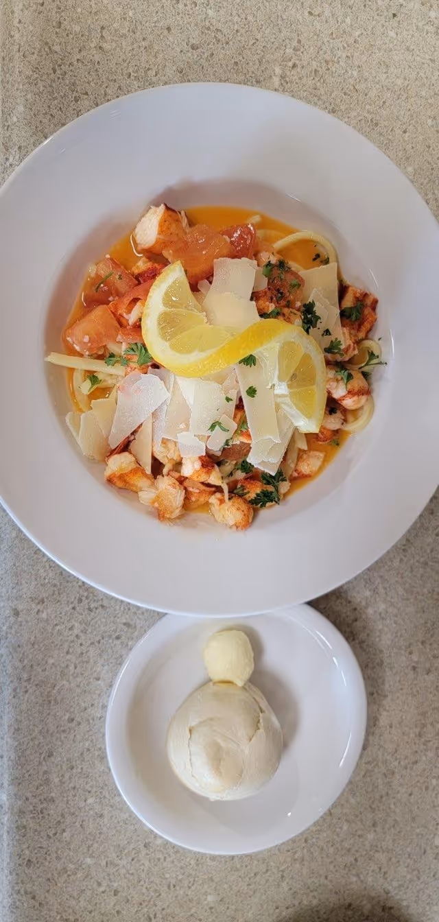A plate of lobster pasta garnished with lemon slices and parmesan cheese, accompanied by a small round bread roll and butter on a white dish.