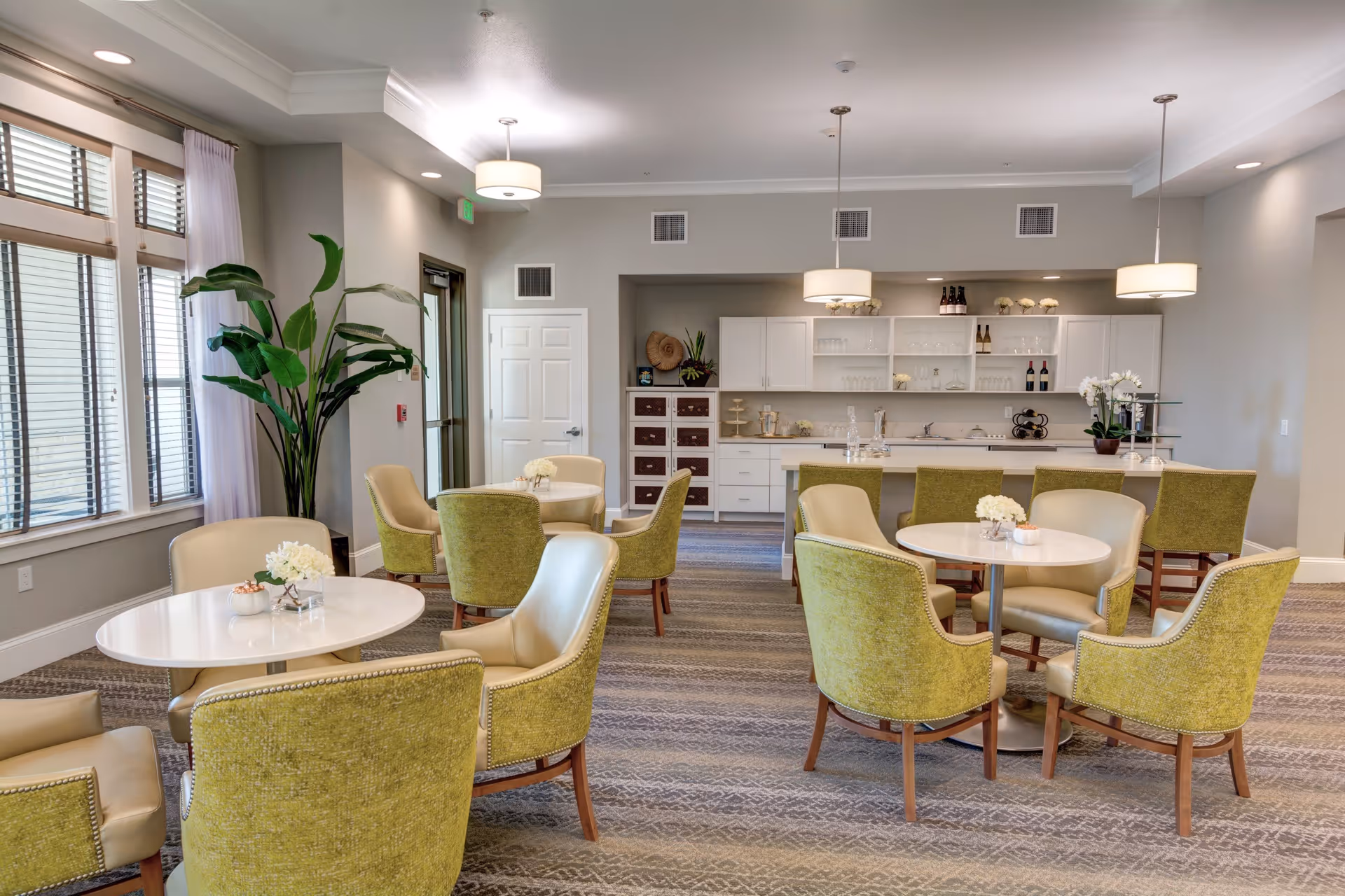 A bright and spacious common area with several round tables surrounded by green and beige upholstered chairs. The room features large windows with white curtains, a tall green plant in the corner, and a kitchenette in the background with white cabinets, shelves, and a countertop. Pendant lights hang from the ceiling, illuminating the space.