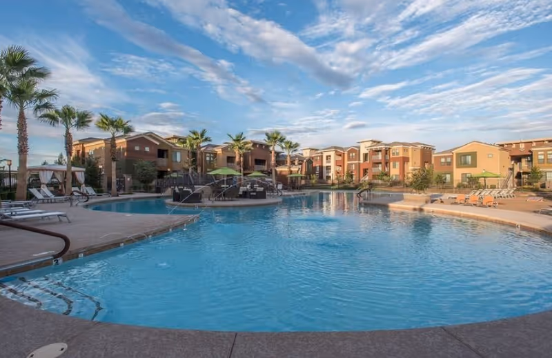 Large outdoor swimming pool with lounge chairs, palm trees, and surrounding apartment-style buildings under a blue sky.