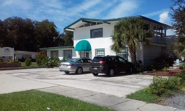 Exterior view of a two-story white building with a green awning over the entrance, surrounded by palm trees and parked cars in the driveway under a partly cloudy sky.