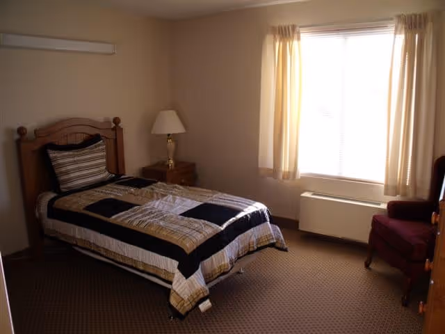 A small bedroom with a single bed covered in a striped and checkered comforter, a wooden headboard, a nightstand with a lamp, a window with beige curtains, a maroon armchair, and a carpeted floor.