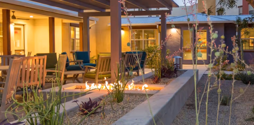 Outdoor courtyard patio with chairs and a rectangular fire pit under a pergola in front of the facility building.