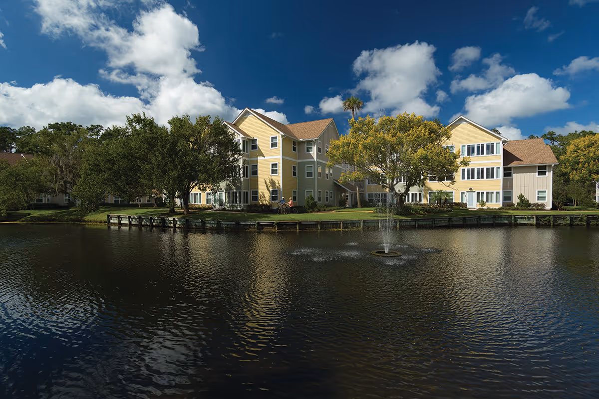 Yellow multi-story building by a pond with a central fountain, trees, and a small dock under a partly cloudy sky.
