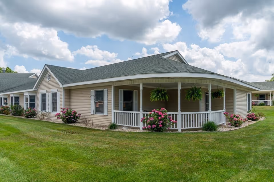 Single-story beige senior living building with a covered wraparound porch, hanging plants, flowering shrubs, and a manicured lawn under a cloudy sky.