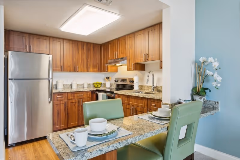 Modern kitchen with wooden cabinets, granite countertops, stainless steel refrigerator and stove, and a breakfast bar with two green chairs set with white dishes and cups. A white orchid plant is placed on the countertop near a light blue wall.