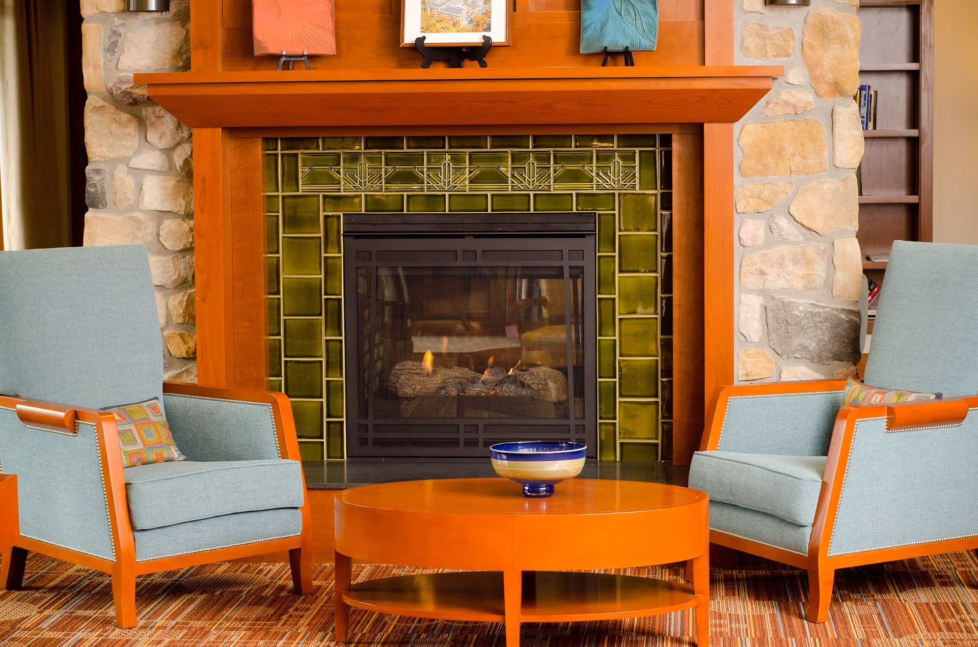 Two upholstered armchairs flank a lit fireplace with a green tile surround and a round wooden coffee table in front.