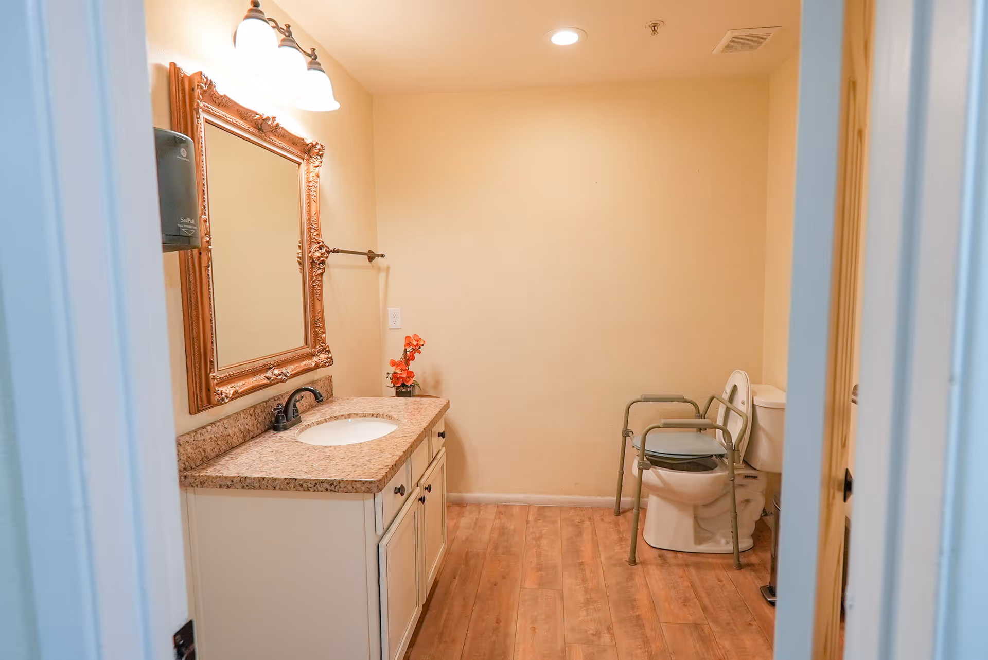 A bathroom with a wooden floor, a white vanity with a granite countertop and an oval sink, a large ornate framed mirror above the sink, a wall-mounted light fixture with two bulbs, a small vase with orange flowers on the countertop, and a toilet equipped with a metal safety frame for assistance.